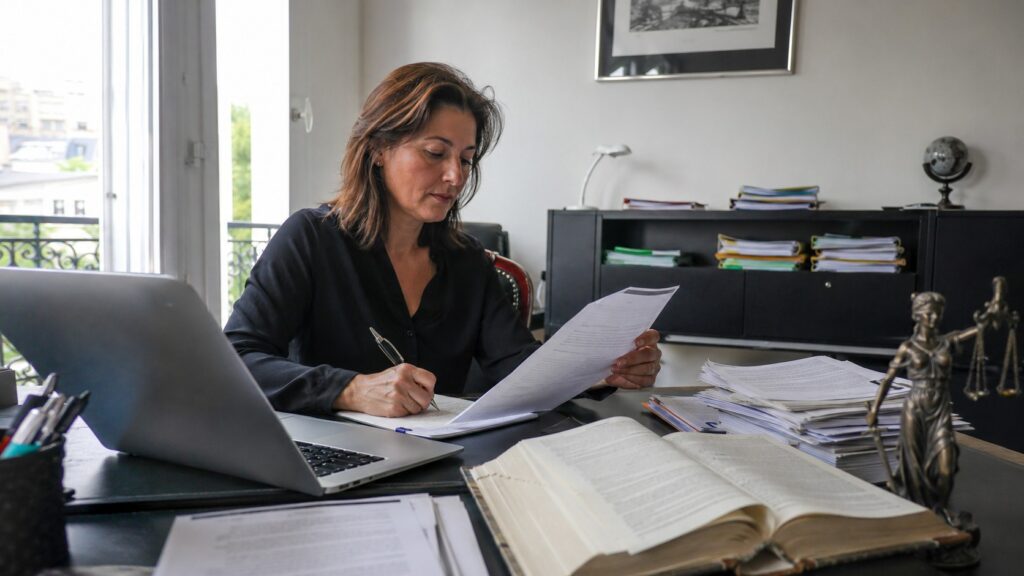 Agence de traduction juridique certifiée ISO 17100, traductrice professionnelle examinant des documents juridiques à un bureau avec ordinateur portable, contrats imprimés, ouvrage de référence et statue de la Justice, photo documentaire Panasonic GH4 grand-angle 24-28 mm, lumière naturelle ensoleillée, juillet 2026