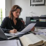 Agence de traduction juridique certifiée ISO 17100, traductrice professionnelle examinant des documents juridiques à un bureau avec ordinateur portable, contrats imprimés, ouvrage de référence et statue de la Justice, photo documentaire Panasonic GH4 grand-angle 24-28 mm, lumière naturelle ensoleillée, juillet 2026