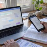 IA de traduction utilisée sur ordinateur portable avec interface conversationnelle générique, comparaison d’un document sur smartphone, dictionnaire ouvert, rapports imprimés et carnet de notes sur bureau en bois près d’une fenêtre, contrôle humain des limites de la traduction automatique, lumière naturelle ensoleillée, prise de vue documentaire Panasonic GH4, grand-angle 24–28 mm, profondeur de champ modérée, couleurs neutres peu saturées, format 16:9, août 2026