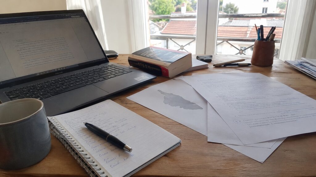 Traduction corse français sur bureau de traducteur avec ordinateur portable, documents de relecture, carnet de notes, stylo, ouvrage linguistique et carte de la Corse, photographie documentaire Panasonic GH4 grand-angle 24-28 mm en lumière naturelle ensoleillée, préparation de contenu avant publication, 2026