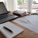 Traduction corse français sur bureau de traducteur avec ordinateur portable, documents de relecture, carnet de notes, stylo, ouvrage linguistique et carte de la Corse, photographie documentaire Panasonic GH4 grand-angle 24-28 mm en lumière naturelle ensoleillée, préparation de contenu avant publication, 2026