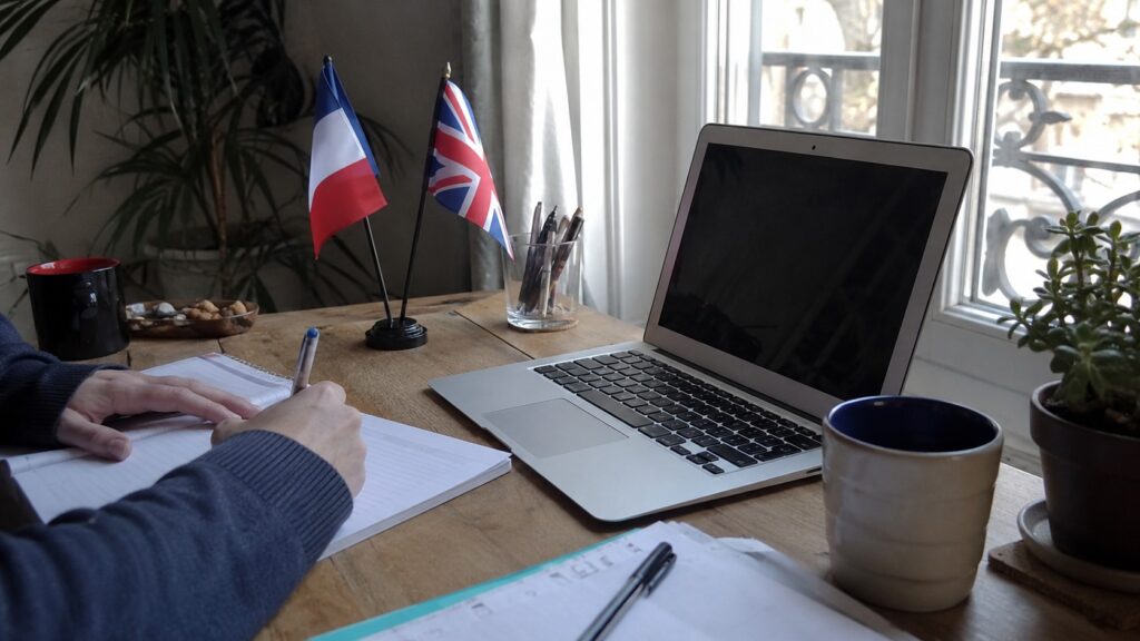 traduction de texte du français vers l’anglais sur ordinateur portable, prise de notes manuscrites dans un cahier, drapeaux français et britannique sur bureau en bois, lumière naturelle près d’une fenêtre, photo documentaire Panasonic GH4 grand-angle 24–28 mm, profondeur de champ modérée, rendu réaliste, juillet 2026