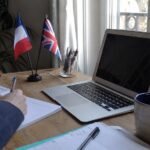 traduction de texte du français vers l’anglais sur ordinateur portable, prise de notes manuscrites dans un cahier, drapeaux français et britannique sur bureau en bois, lumière naturelle près d’une fenêtre, photo documentaire Panasonic GH4 grand-angle 24–28 mm, profondeur de champ modérée, rendu réaliste, juillet 2026
