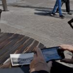 traduction d'une photo avec smartphone au café en extérieur, personne photographiant une image imprimée noir et blanc posée sur table en bois, prise de vue documentaire au Panasonic GH4, grand-angle 24-28 mm, lumière naturelle ensoleillée, profondeur de champ modérée, textures réalistes, juillet 2026