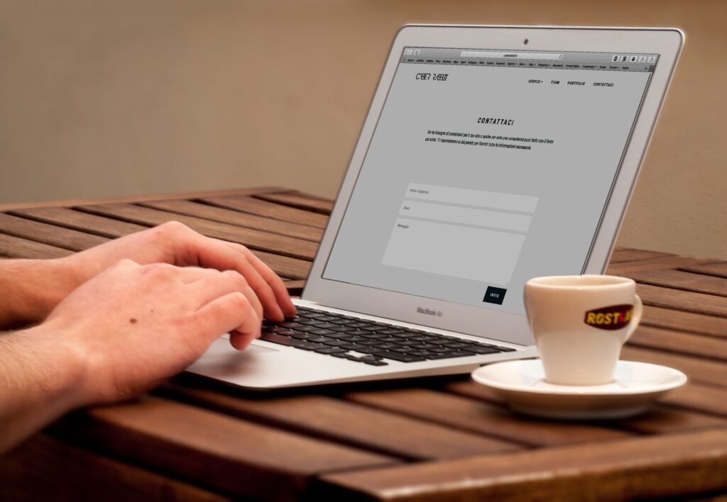 Utilisateur saisissant un message dans le formulaire de contact d’un site web affiché sur un MacBook Air, mains sur le clavier, tasse de café expresso et soucoupe posées sur une table en bois, travail en ligne, 2026, format 16:9 JPG