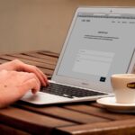 Utilisateur saisissant un message dans le formulaire de contact d’un site web affiché sur un MacBook Air, mains sur le clavier, tasse de café expresso et soucoupe posées sur une table en bois, travail en ligne, 2026, format 16:9 JPG