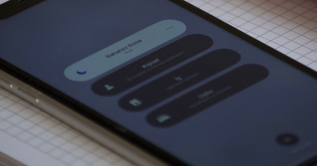 Gros plan sur un smartphone posé en diagonale sur un cahier quadrillé, application de traduction vocale ouverte avec champ de saisie, bouton Traduire, icônes microphone et appareil photo, carnet rouge à pages jaunies contenant des notes manuscrites en arrière-plan, ordinateur portable partiellement visible, bureau en bois, lumière intérieure douce, faible profondeur de champ, mise au point sur l’écran du téléphone, cadrage vertical rapproché, tons neutres et légèrement chauds, texture réaliste du papier et des appareils, 5 août 2026
