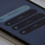 Gros plan sur un smartphone posé en diagonale sur un cahier quadrillé, application de traduction vocale ouverte avec champ de saisie, bouton Traduire, icônes microphone et appareil photo, carnet rouge à pages jaunies contenant des notes manuscrites en arrière-plan, ordinateur portable partiellement visible, bureau en bois, lumière intérieure douce, faible profondeur de champ, mise au point sur l’écran du téléphone, cadrage vertical rapproché, tons neutres et légèrement chauds, texture réaliste du papier et des appareils, 5 août 2026