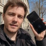 homme testant une application de traduction vocale sur smartphone noir avec interface micro et forme d’onde audio, selfie en extérieur sur un quai parisien au bord de la Seine, immeuble haussmannien en pierre, pont urbain, arbres sans feuilles, ciel bleu et lumière naturelle ensoleillée, cadrage grand-angle 24–28 mm, profondeur de champ modérée, rendu documentaire réaliste Canon EOS R6, août 2026