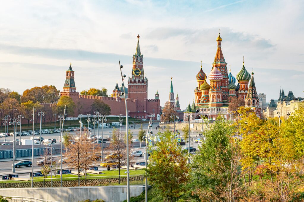 Vue panoramique de Moscou, Kremlin et remparts en briques rouges, tour Spasskaïa avec horloge et étoile, cathédrale Saint-Basile-le-Bienheureux aux coupoles colorées, abords de la place Rouge, circulation automobile, lampadaires urbains, arbres aux feuillages jaunes et orangés d’automne, lumière naturelle ensoleillée, ciel bleu partiellement nuageux, cadrage horizontal grand-angle, profondeur de champ étendue, détails architecturaux nets, couleurs lumineuses, format paysage 16:10, 5 août 2026