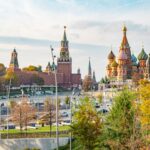 Vue panoramique de Moscou, Kremlin et remparts en briques rouges, tour Spasskaïa avec horloge et étoile, cathédrale Saint-Basile-le-Bienheureux aux coupoles colorées, abords de la place Rouge, circulation automobile, lampadaires urbains, arbres aux feuillages jaunes et orangés d’automne, lumière naturelle ensoleillée, ciel bleu partiellement nuageux, cadrage horizontal grand-angle, profondeur de champ étendue, détails architecturaux nets, couleurs lumineuses, format paysage 16:10, 5 août 2026