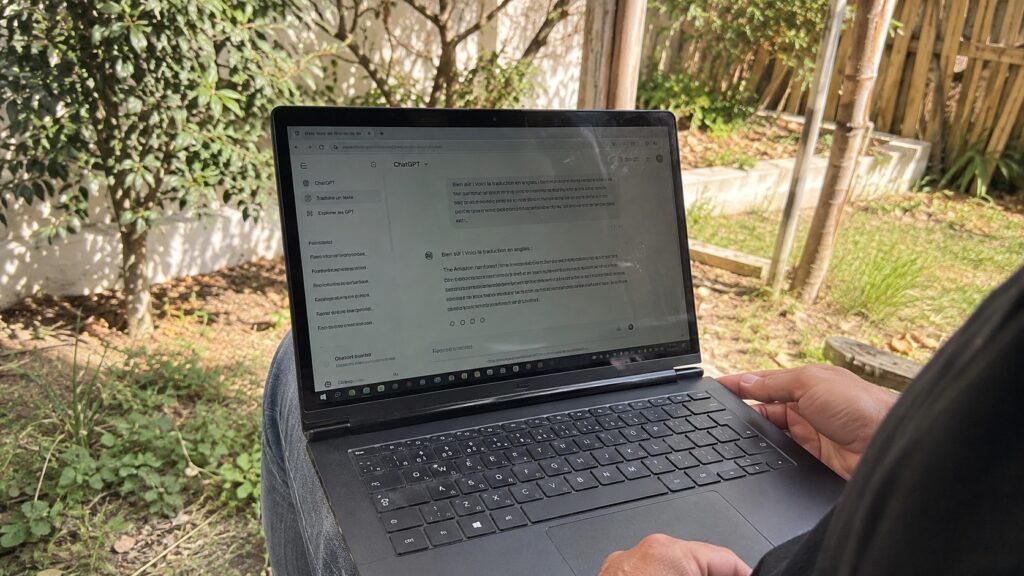 ordinateur portable noir posé sur les genoux d’un utilisateur dans un jardin ensoleillé, interface ChatGPT ouverte avec traduction d’un texte du français vers l’anglais, écran légèrement réfléchissant, clavier AZERTY, mains visibles, végétation et clôture en bois en arrière-plan, prise de vue documentaire au Canon EOS R6, focale grand-angle 24–28 mm, profondeur de champ modérée, lumière naturelle, balance des blancs neutre, textures réalistes, format horizontal 16:9, août 2026