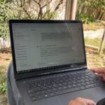 ordinateur portable noir posé sur les genoux d’un utilisateur dans un jardin ensoleillé, interface ChatGPT ouverte avec traduction d’un texte du français vers l’anglais, écran légèrement réfléchissant, clavier AZERTY, mains visibles, végétation et clôture en bois en arrière-plan, prise de vue documentaire au Canon EOS R6, focale grand-angle 24–28 mm, profondeur de champ modérée, lumière naturelle, balance des blancs neutre, textures réalistes, format horizontal 16:9, août 2026